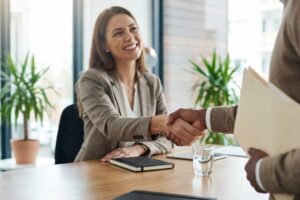 Two professionals shake hands across a wooden table in a bright office, smiling during a meeting.
