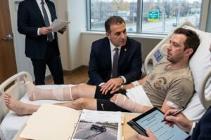 Hospital room scene: patient with leg casts in bed while two suited men discuss and review papers and a tablet nearby.